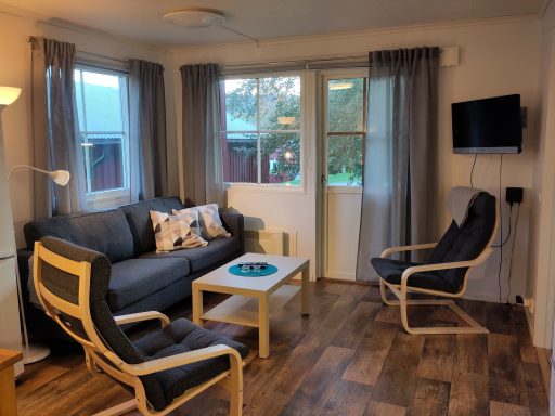 Mysig vardagsrum med soffa, fåtöljer och bord, fönster med utsikt och TV på väggen.