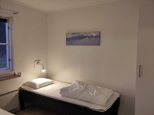 En enkel säng i ett rum med vit vägg och en tavla av snöiga berg.