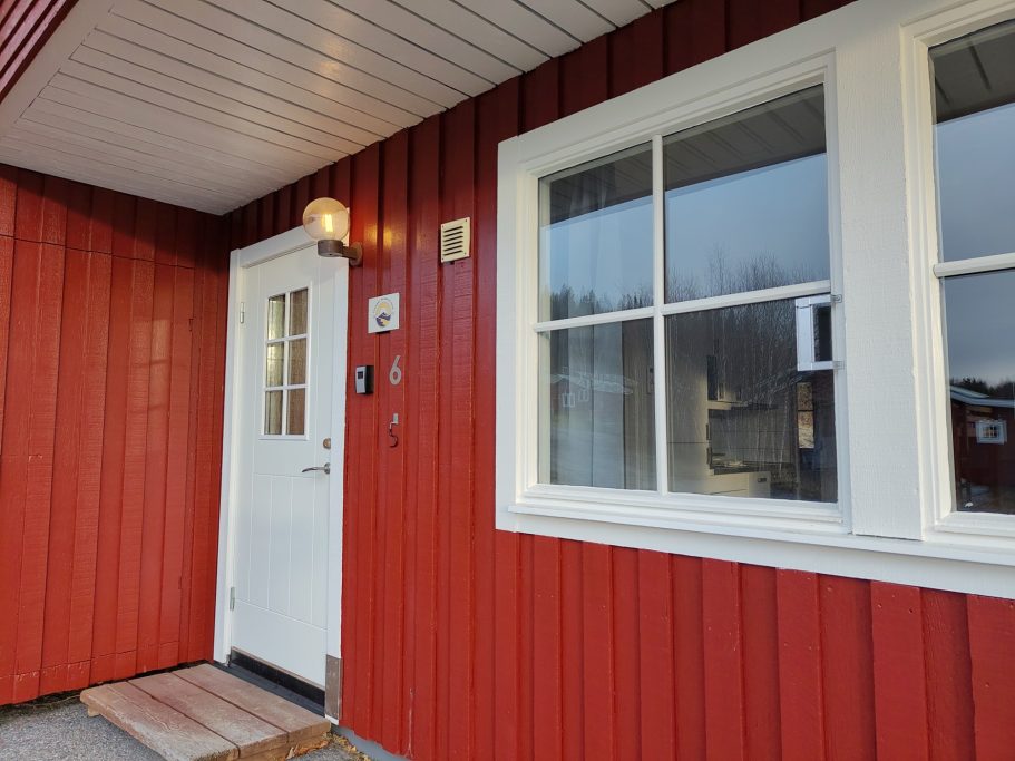 Entré stuga 6 Röd stuga med vit dörr och fönster, belägen i ett lugnt område.