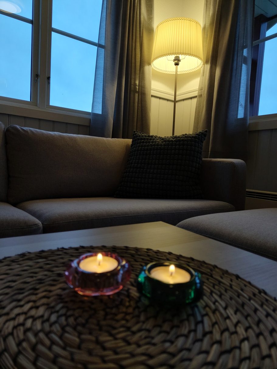 Stugmys Två tända ljus i skålar på ett bord, med en fåtölj och lampa i bakgrunden.