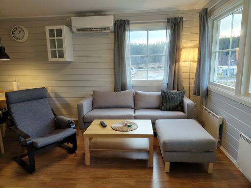 Vardagsrum med en grå soffa, fåtölj, bord och fönster som släpper in ljus.