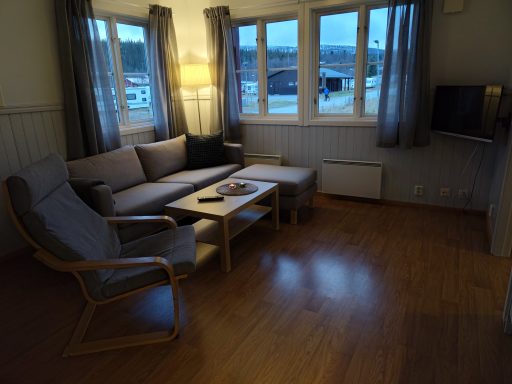 Ljust vardagsrum med soffa, fåtölj, bord och fönster med gardiner.