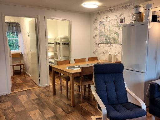 Ljust kök med matbord, stolar, kylskåp och en fåtölj, tapet med blommönster.