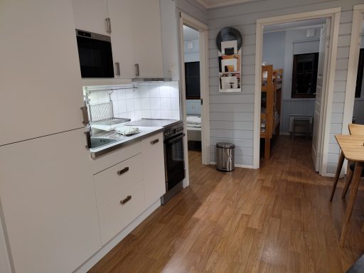 Ljust kök med moderna vitvaror och trägolv, öppet mot ett vardagsrum.