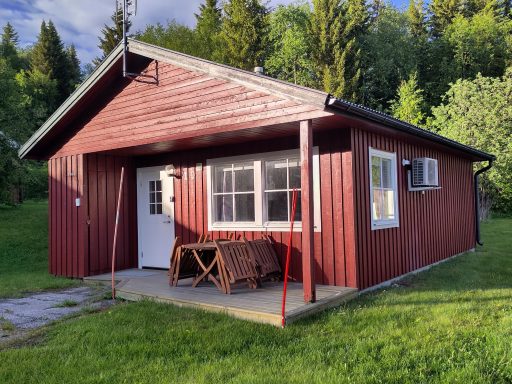 Röd stuga med vita fönster, omgiven av grönska och träd.