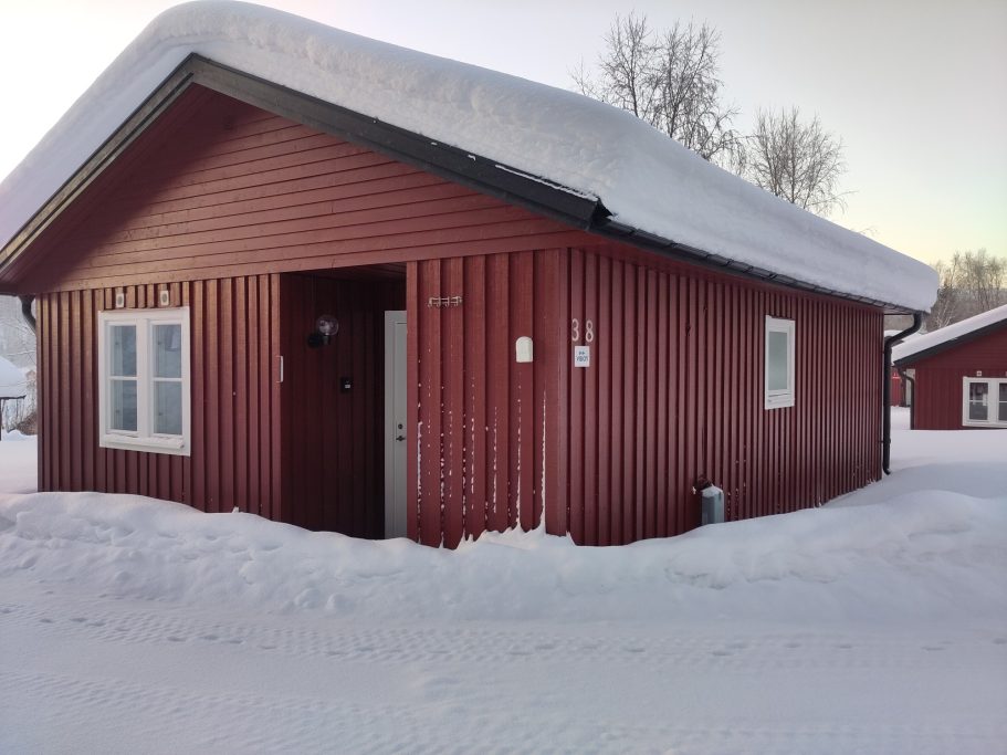 Stuga 38 Röd stuga med snö på taket, omgiven av snöiga marker.