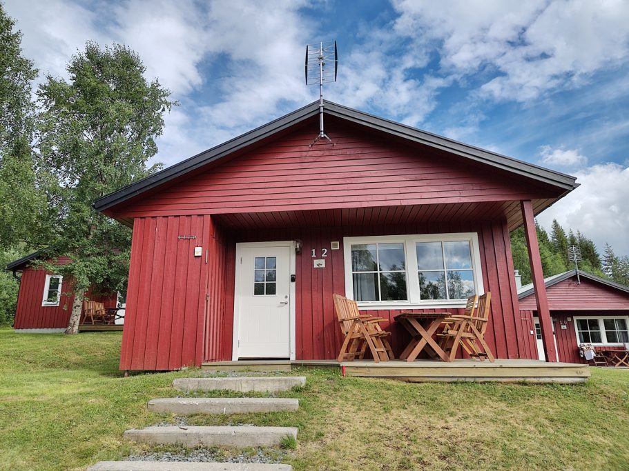 Stuga 12 Röd stuga med terrasse och utsikt under en blå himmel med vita moln.