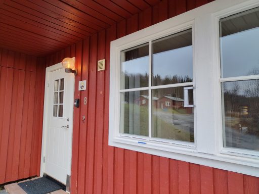 Röd stuga med vit dörr och fönster, delvis mulet väder i bakgrunden.