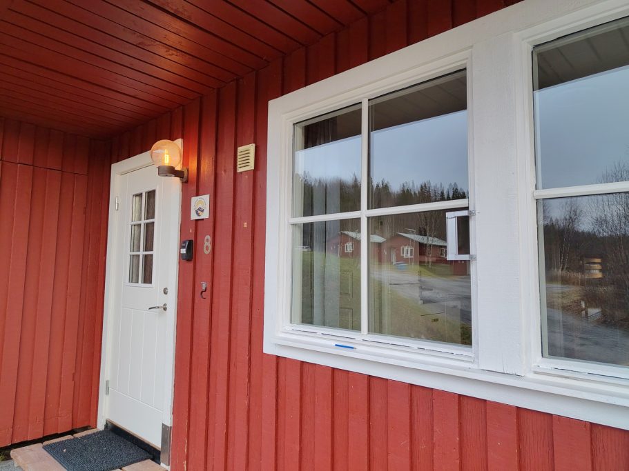 Entré Röd stuga med vit dörr och fönster, delvis mulet väder i bakgrunden.