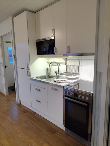 Kök med vit inredning, spis, mikrovågsugn och diskho under belysning.