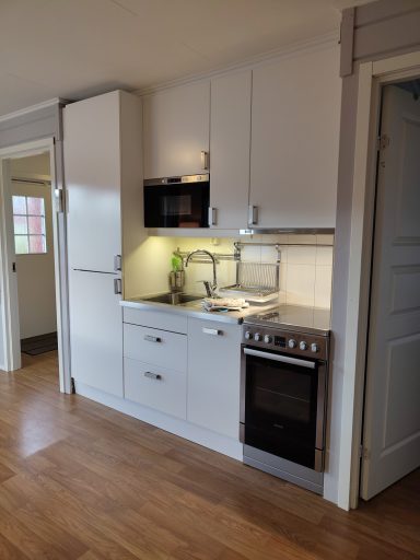 Ljust kök med vit inredning, ugn, spis, mikrovågsugn och träplankor på golvet.