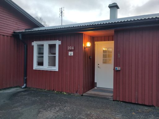 Röd stuga med vit dörr och fönster, belyst av en lampa vid entrén.