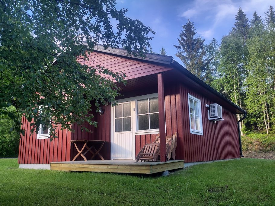 Stuga 38 Röd stuga med verandagolv omgiven av grönska och träd.