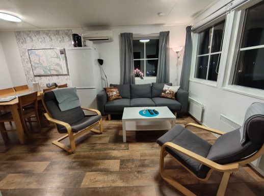 Modernt vardagsrum med soffa, fåtöljer och matbord, ljus inredning och mörka gardiner.