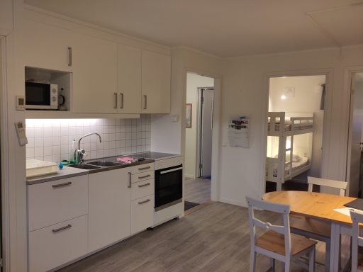 Köksutrymme med vit inredning, matbord och dörr till hall med våningssäng.