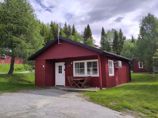 Röd stuga med veranda omgiven av träd och grön gräsmatta.