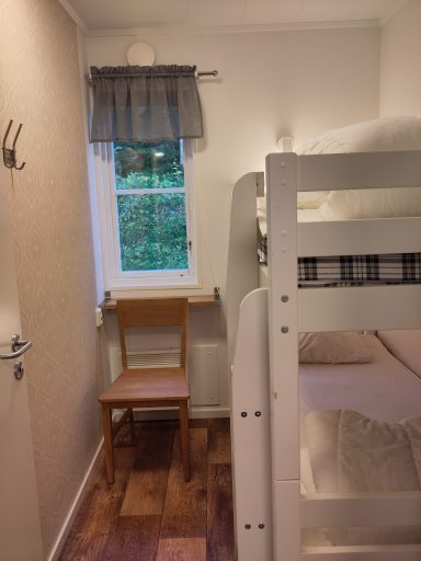 Litet rum med våningssäng, stol och fönster med gardin.