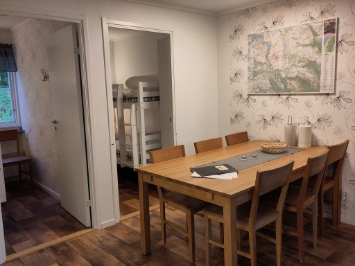 Ljust rum med träbord, stolar och en väggdekoration av kartor. Bunkbeds i bakgrunden.